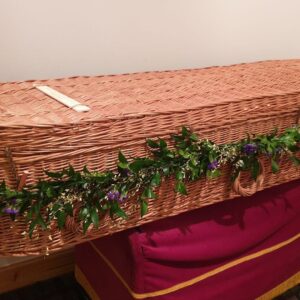 Wicker coffin with lush green and purple floral spray